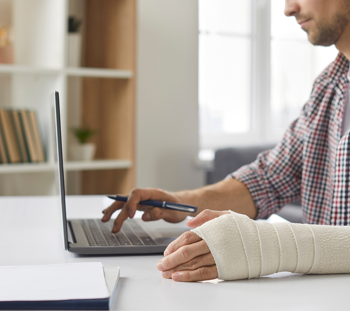 A person with a plaid shirt works on a laptop at a desk. Their right hand is in a cast, holding a pen. The setting is a bright, modern office space.